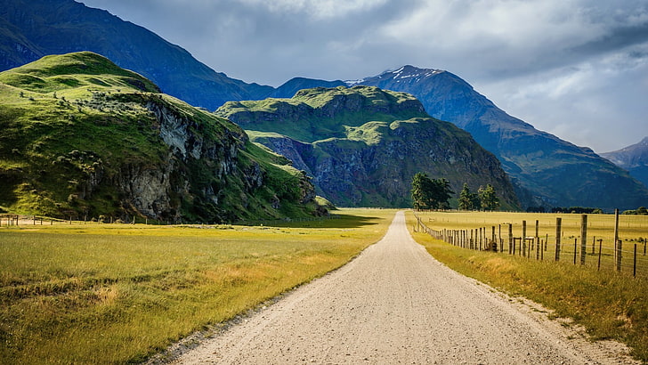 طريق وعرة رمادية بجانب الحقول الخضراء عبر الجبال والمناظر الطبيعية والطرق والجبال والطبيعة، خلفية HD
