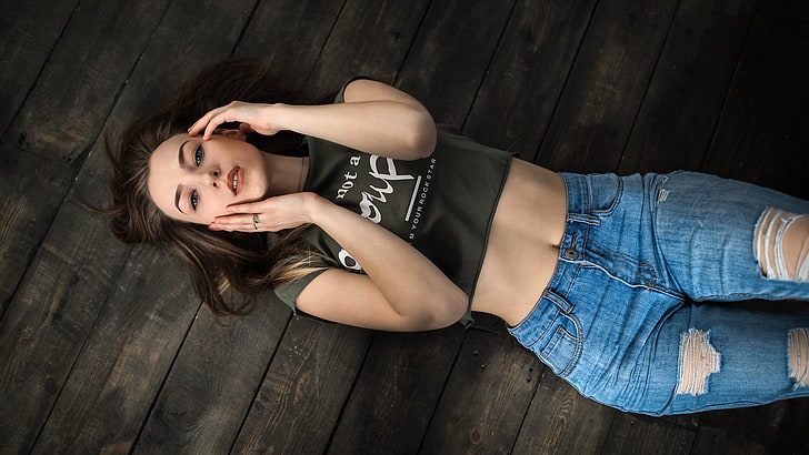 women's black crew-neck crop top, women, top view, brunette, torn jeans, blue eyes, lying on back, belly, wooden surface, portrait, touching face, short tops, looking at viewer, HD wallpaper