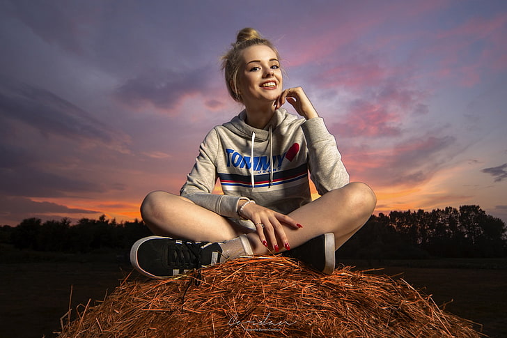 women's gray and blue pullover hoodie, women, model, looking at viewer, blonde, hairbun, smiling, portrait, sunset, sneakers, red nails, sitting, legs crossed, straw, sweatshirts, women outdoors, hay, HD wallpaper