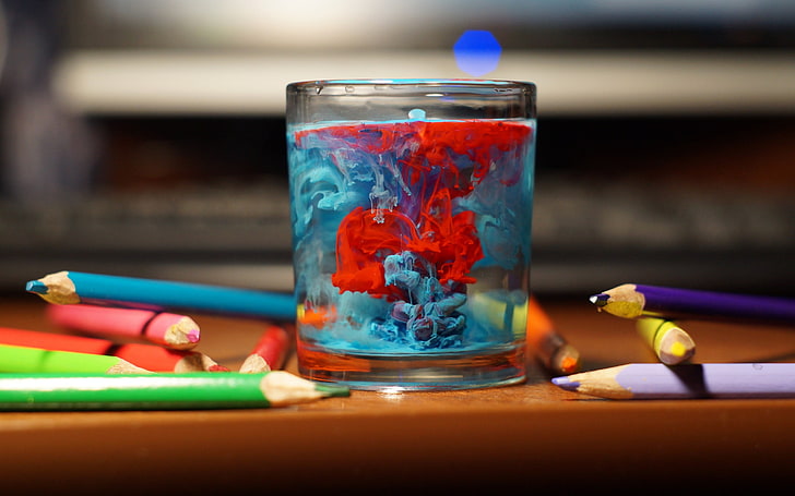прозрачна стъклена чаша и писалки от различни цветове, накланяне на фокус, фотография на чаша за пиене с червен и син цвят вътре в декора, маса, чаша, вода, моливи, пръски боя, цветни, дълбочина на рязкост, фотография, боке, боя във вода, акварел, HD тапет