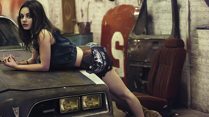 Mila Kunis, Frau, die sich auf ein schwarzes Pontiac-Auto stützt, Mila Kunis, Frauen, Schauspielerin, Frauen mit Autos, Brünette, Garagen, Beige, gebeugt, Minirock, HD-Hintergrundbild